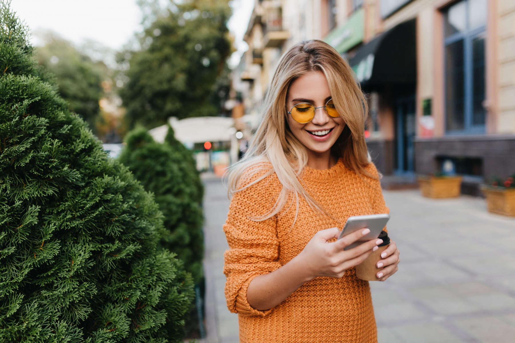 lovely-blonde-woman-walking-by-green-bushes-with-smile-carrying-smartphone-cup-coffee.jpg